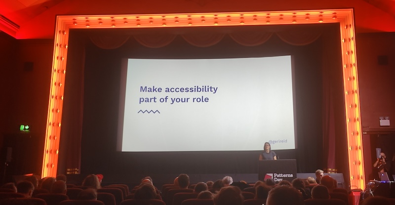 On the stage of a cinema is a speaker and their slide deck projected on the screen with the text, Make accessibility part of your role