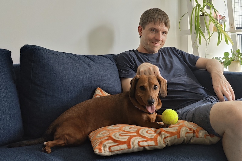 Calum sitting on a blue sofa with Coco the dog panting, looking intently at a tennis ball