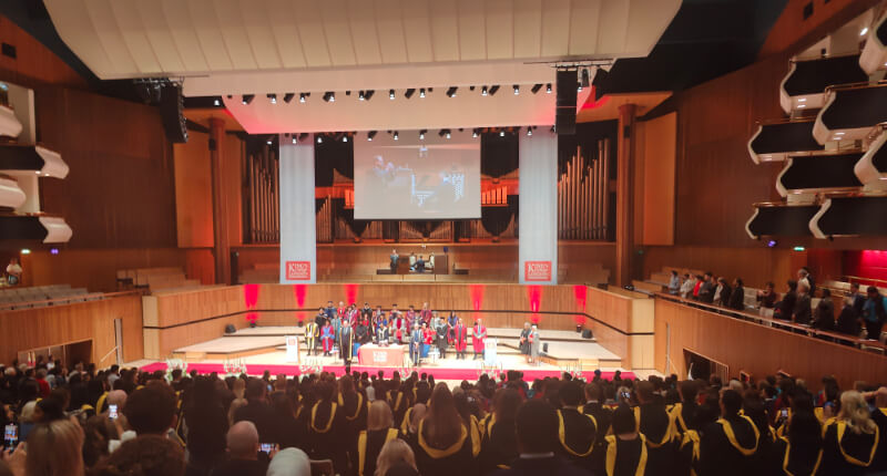 Graduation ceremony inside Royal Festival Hall looking down on the stage with many people dressed in black graduation gowns