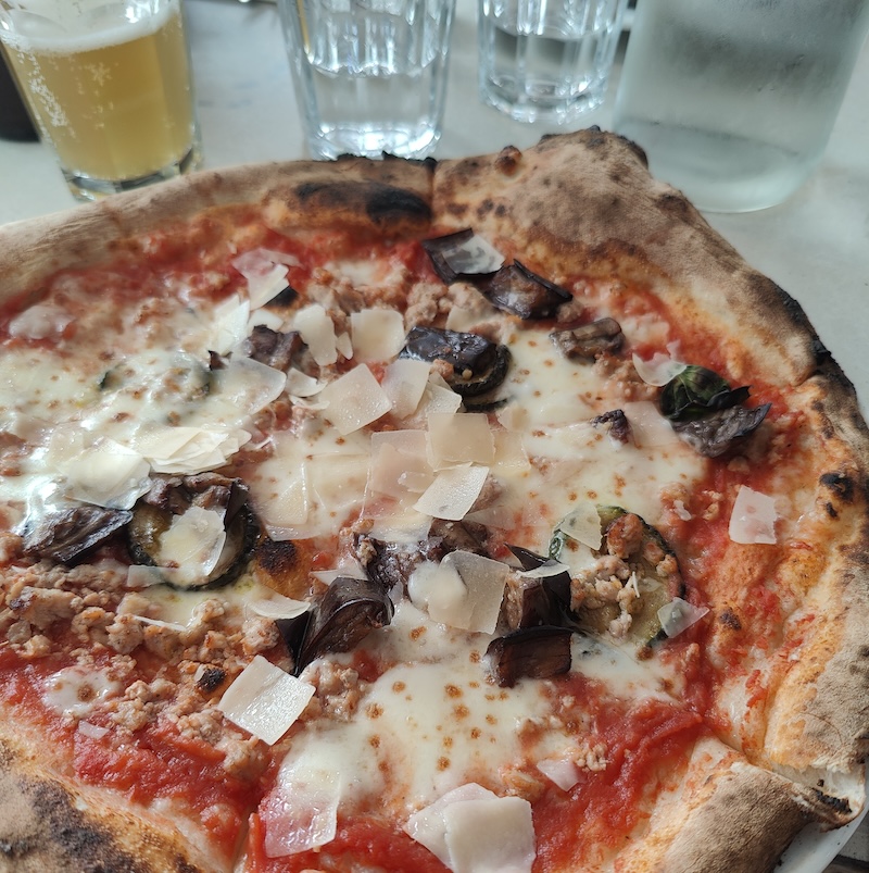 Close-up shot of pizza with shavings of Parmesan and aubergine pieces and a light-coloured ale above