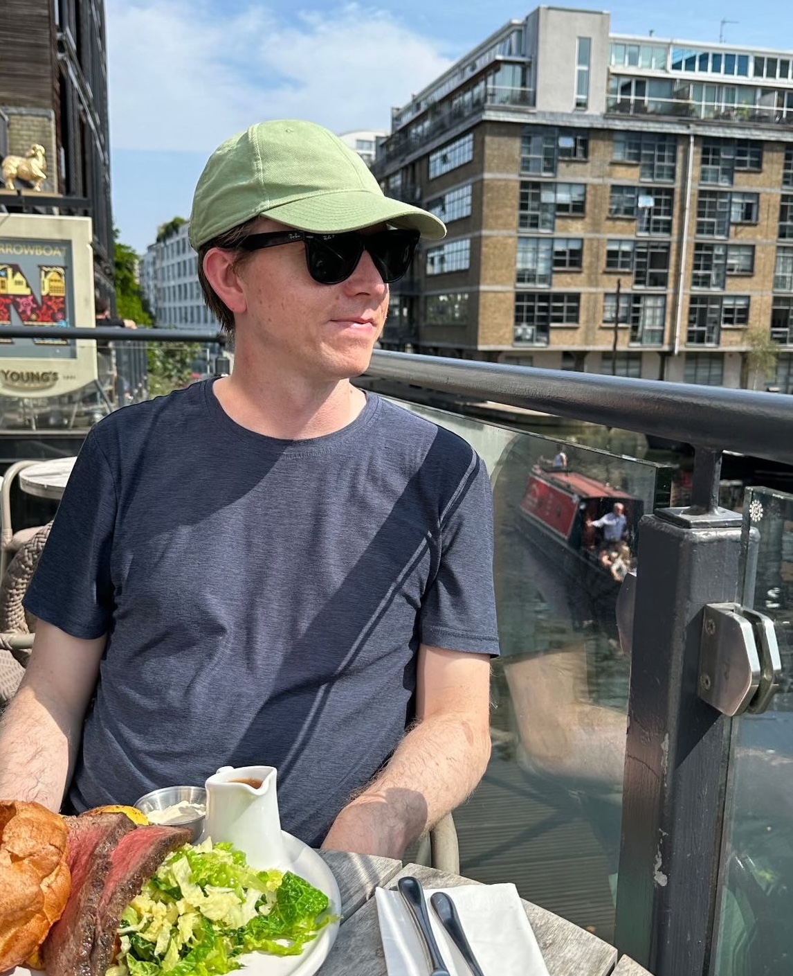 Calum wearing a green cap and sunglasses sits at an outdoor restaurant table by a canal, with buildings and a canal boat in the background