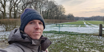 Calum beside a green gate in tree-lined countryside with some snow on the field
