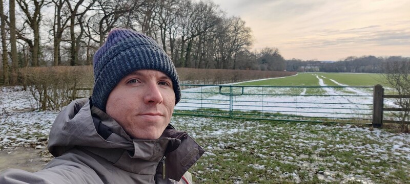 Calum beside a green gate in tree-lined countryside with some snow on the field
