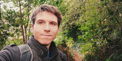 Calum in a grey coat with backdrop of tree-line footpath