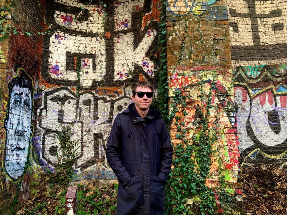 Calum in long coat with backdrop of graffiti on railway architecture