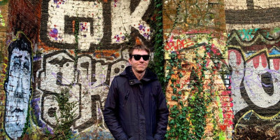 Calum in long coat with backdrop of graffiti on railway architecture