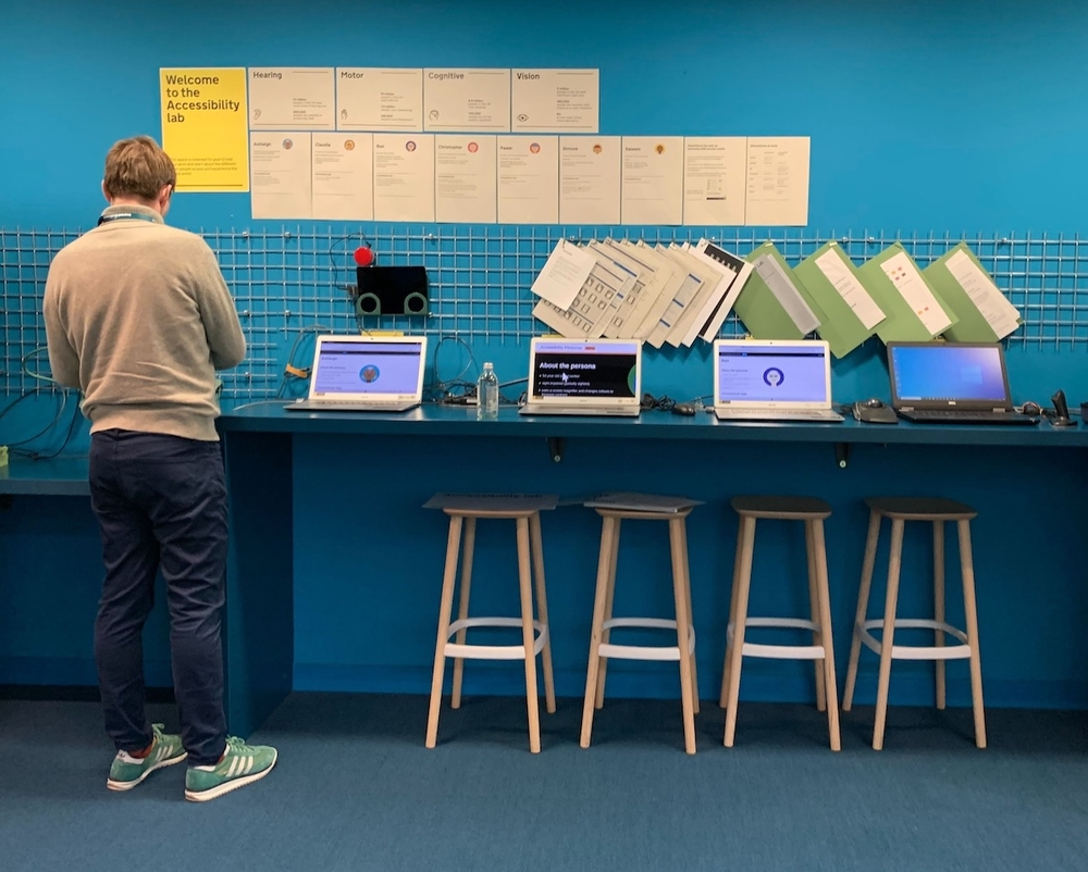 man stands at a blue counter displaying multiple laptops, and accessibility personas described on posters above