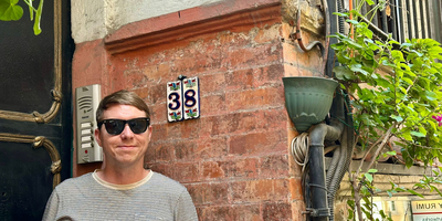 The house number 38 on ceramic tiles with a pretty backdrop of plants and brick wall, Calum stand beside wearing sunglasses