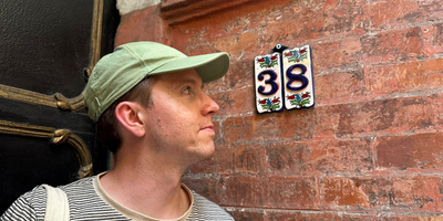 The house number 38 on ceramic tiles with a pretty backdrop of plants and brick wall, Calum stands beside looking at the number